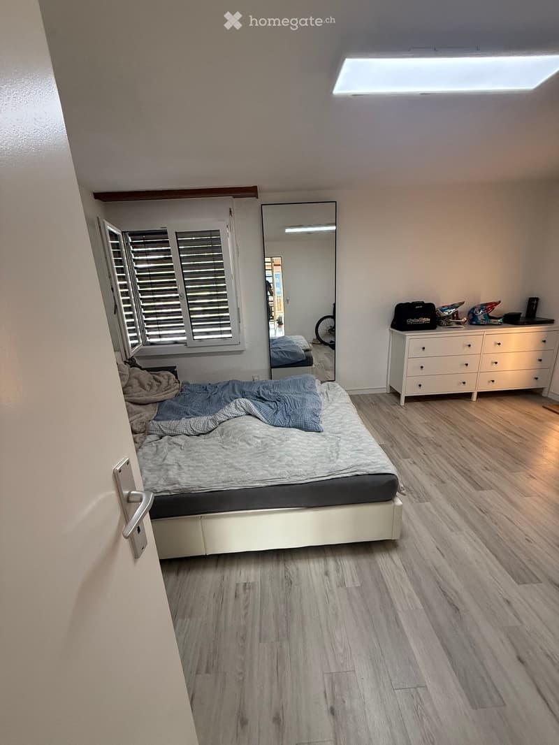 A modern bedroom with a white bed, wooden flooring, a large mirror, and a dresser with various items on top.