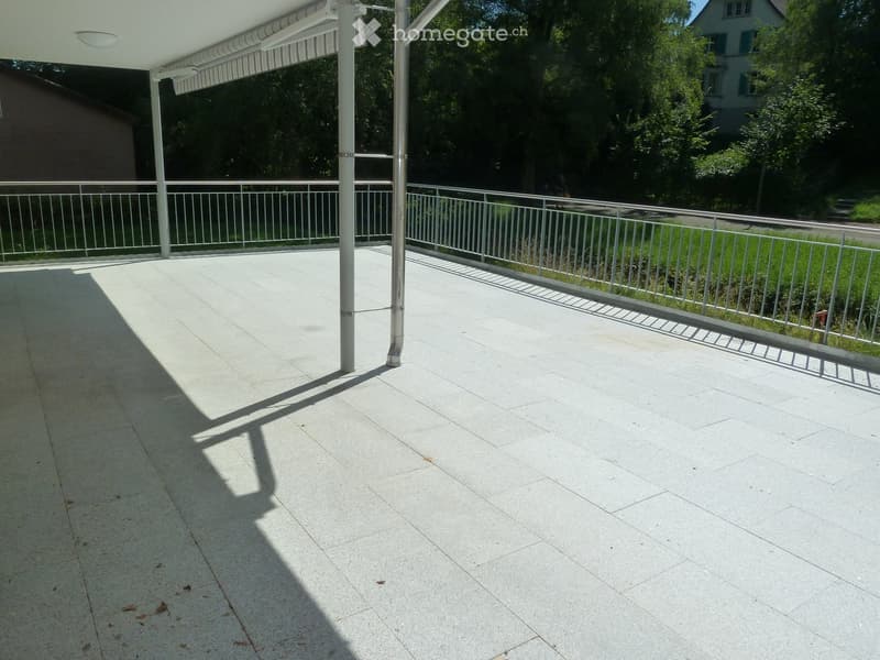 A covered balcony with a tiled floor, metal railing, and a retractable awning, overlooking a green area with trees and a neighboring house.