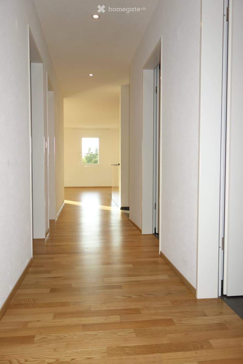 Bright hallway with wooden flooring, white walls, and recessed lighting. Features multiple doors and a window at the far end.