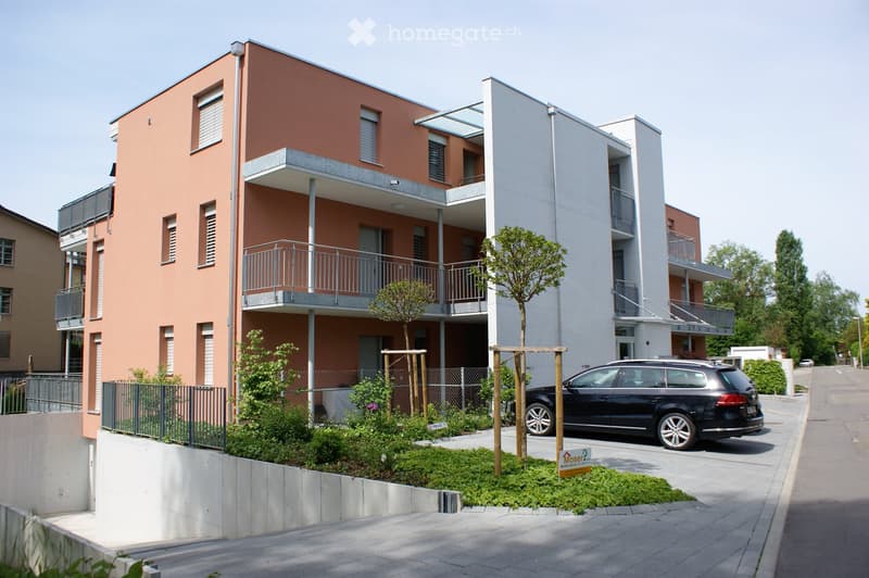 Modern two-story residential building with orange and white facade, balconies, and a small garden area in front.
