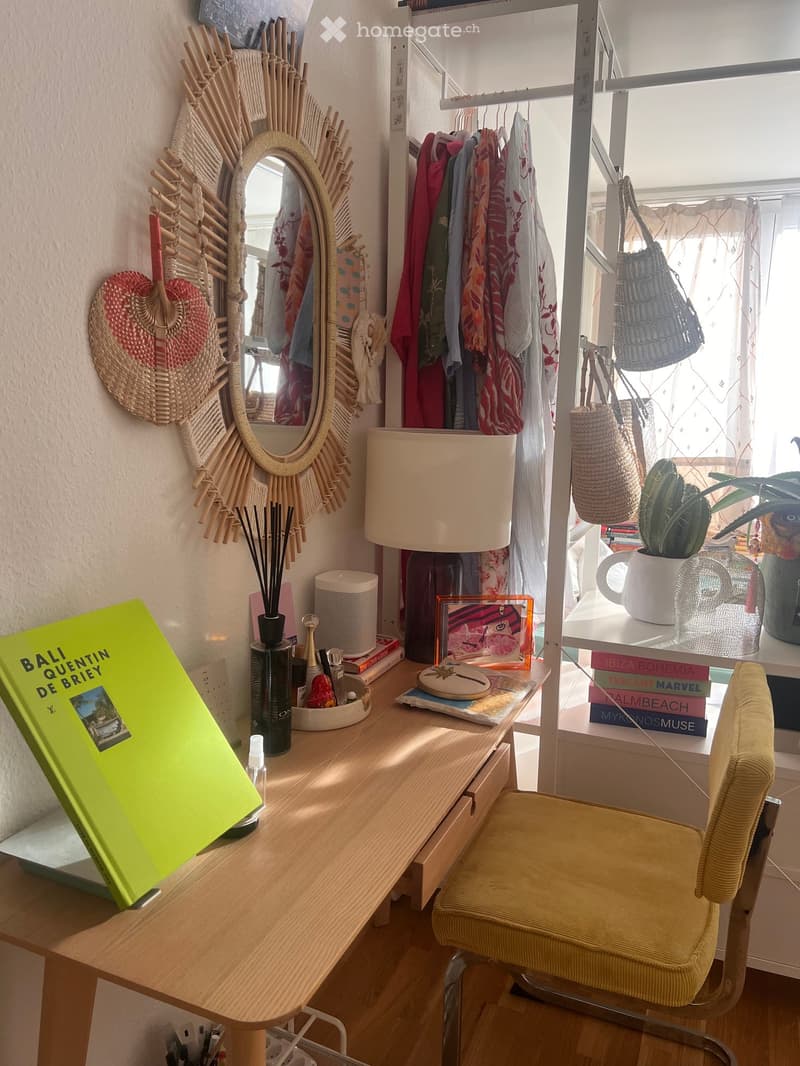 A cozy, well-lit room with a wooden desk, a yellow chair, a round mirror with a woven frame, a lamp, books, a plant, and decorative items.