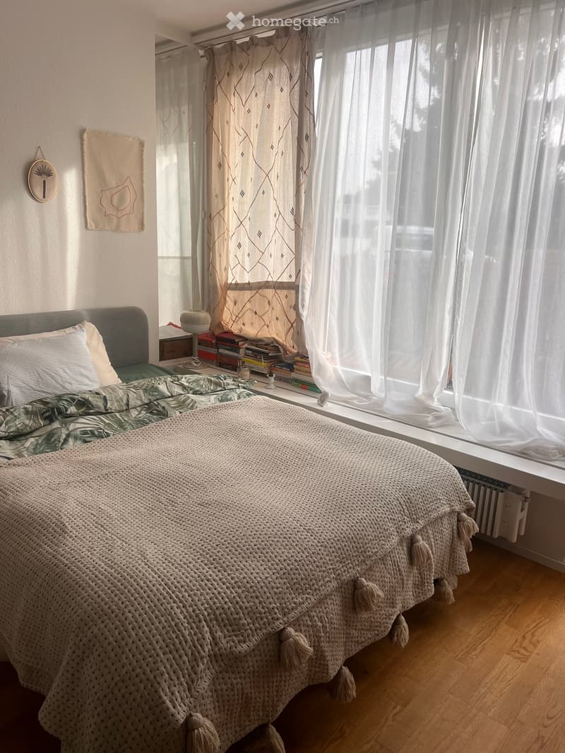 Cozy bedroom with a large bed covered in a textured blanket and pillows, a bedside table with a lamp, a stack of books, and decorative wall hangings near a large window with sheer curtains.