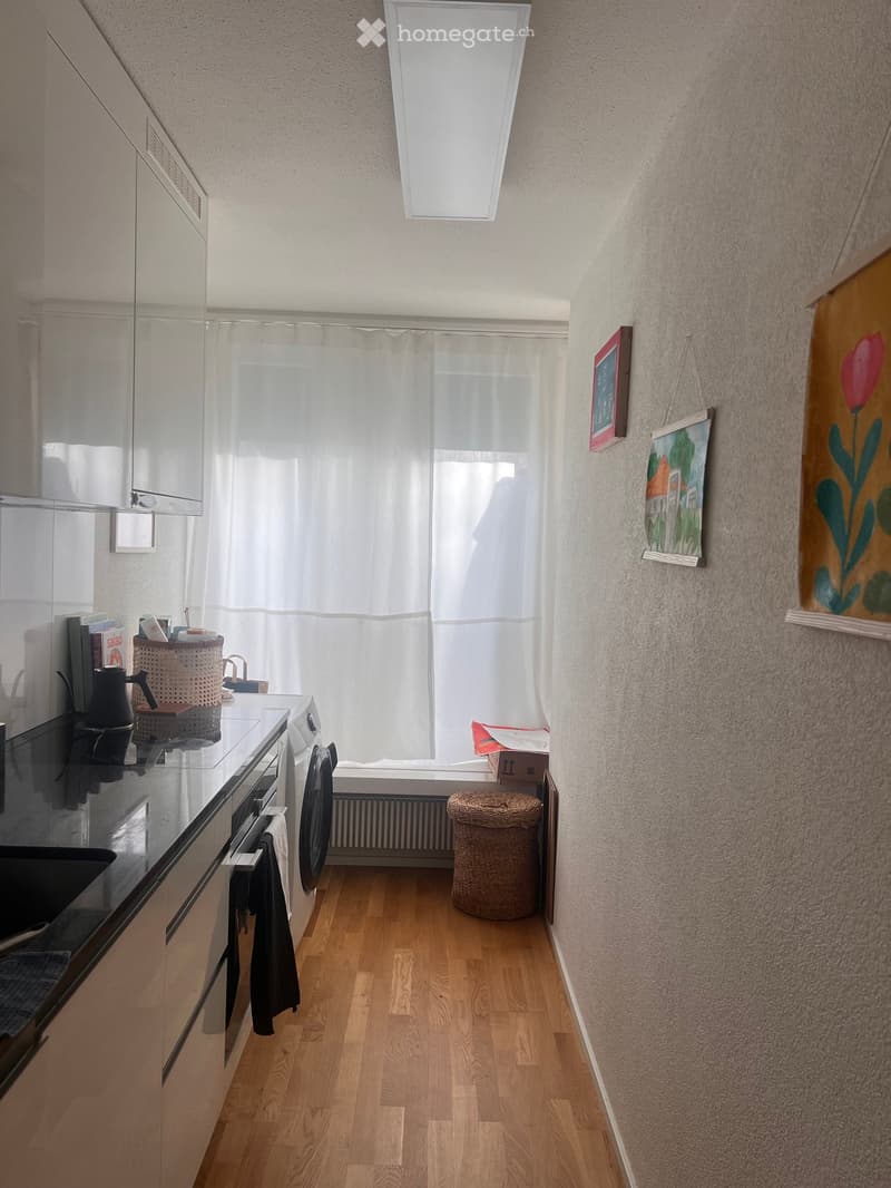 Bathroom with wooden flooring, white walls, a sink with a mirror, a washing machine, a small stool, and a window with a curtain. There are books and a basket on the sink counter.