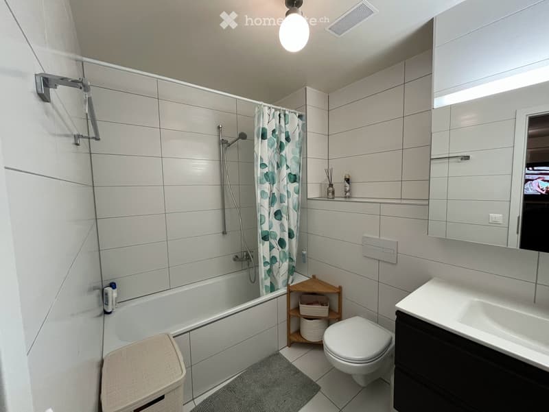 Modern bathroom with a bathtub, toilet, sink, mirror, and a wooden shelf holding baskets. The shower area has a curtain with a leafy pattern, and the walls are tiled in light gray.