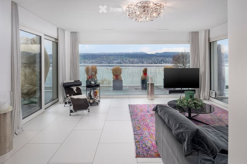Modern living room with large windows offering a scenic view, featuring a grey sofa, coffee table, armchair, television, and decorative items.