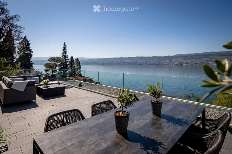Balcony with a view of a lake, featuring a sofa, coffee table, wicker chairs, and potted plants.