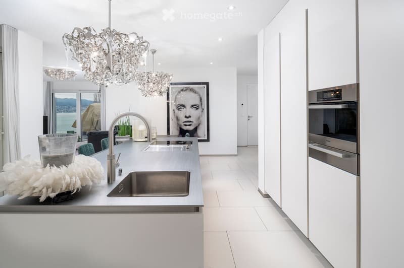 Modern kitchen with stainless steel sink, faucet, built-in oven, and chandelier. Features a large window with a scenic view, a dining area with a table and chairs, and decorative items on the counter.