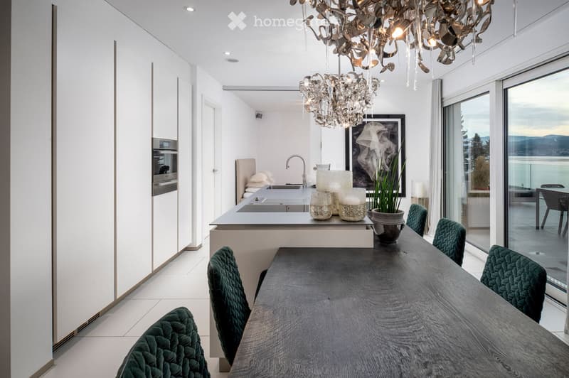 Modern kitchen and dining area with a sleek island, dark wooden dining table, green upholstered chairs, and a chandelier. The kitchen features white cabinets, a stainless steel oven, and a large window overlooking a scenic view.