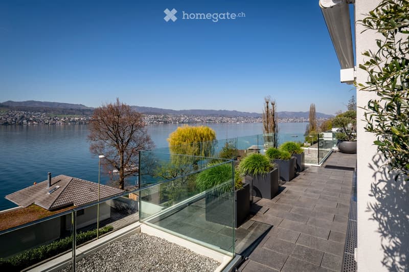 A modern balcony with a glass railing, overlooking a scenic lake and distant mountains. The balcony features potted plants and a paved surface.