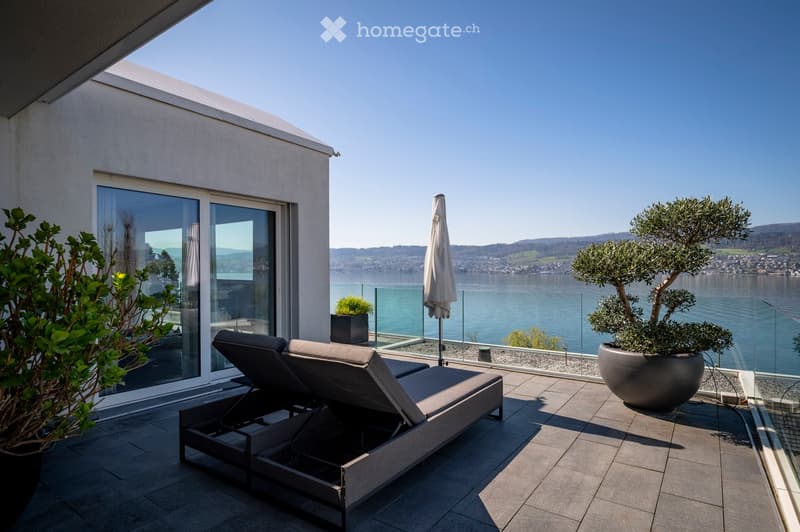 Modern rooftop terrace with sun loungers, umbrella, and potted plants, overlooking a scenic lake and distant hills.