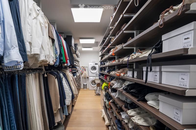 Well-organized walk-in closet with wooden shelves and clothing racks, featuring neatly arranged clothing, shoes, and accessories, with a washing machine in the background.