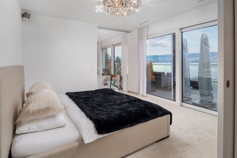 Modern bedroom with large windows offering a scenic view, a large bed with pillows and a fur throw, a decorative toy horse, and a chandelier.