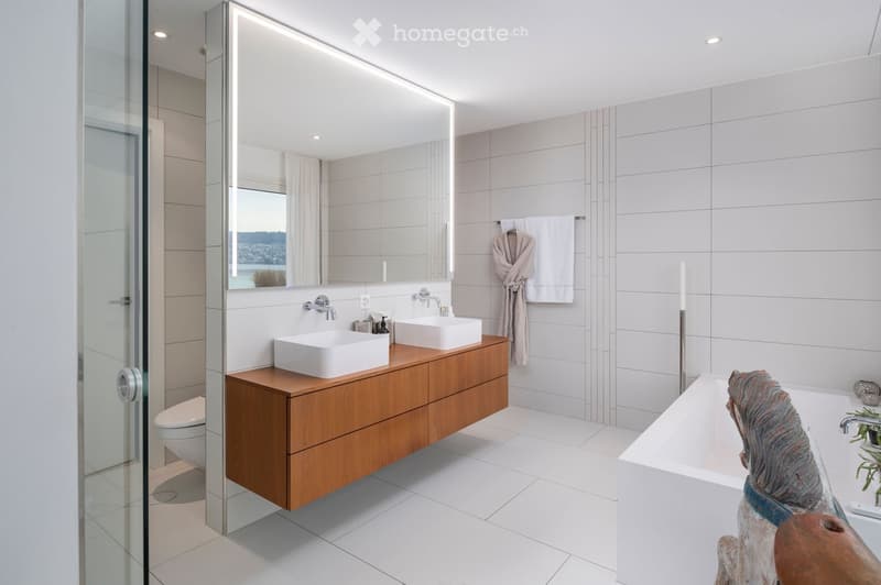 Modern bathroom with white tiles, wooden vanity, double sinks, large mirror, bathtub, and a view through a glass door.