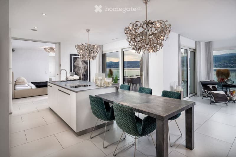 Modern open-plan kitchen and dining area with a central island, sleek dining table, green upholstered chairs, and a chandelier. Large windows offer a scenic view of the landscape.