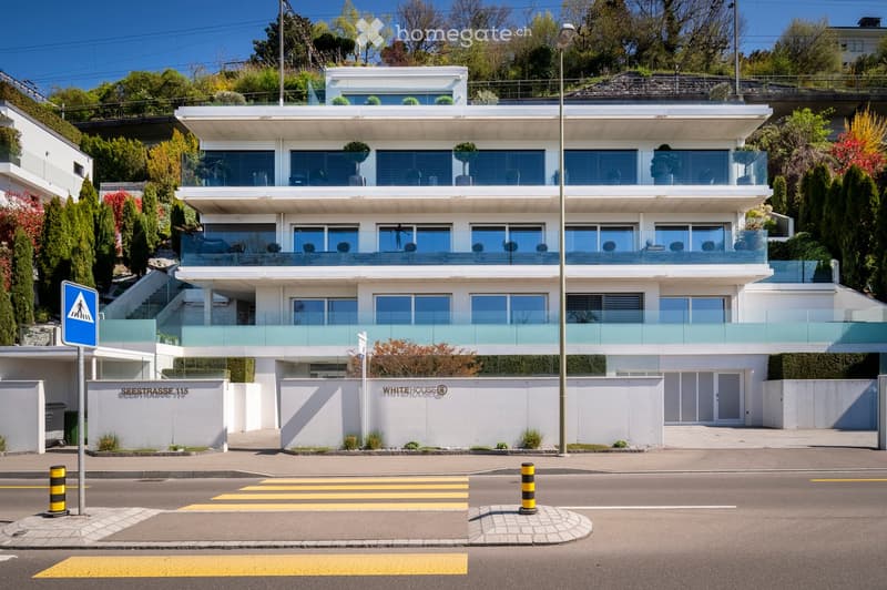 Modern, multi-story white building with large windows and balconies, situated on a hillside with surrounding greenery.