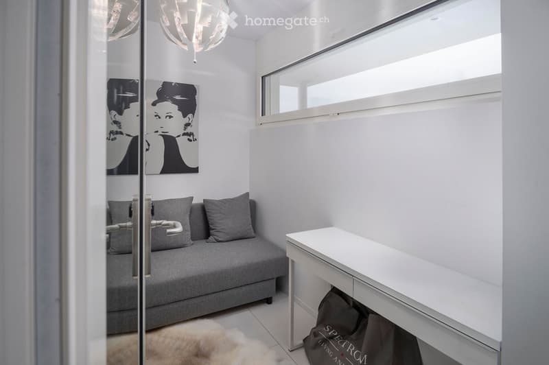 Modern, compact living space with a grey sofa, white desk, and a large window letting in natural light. A picture frame and a fur rug are also visible.