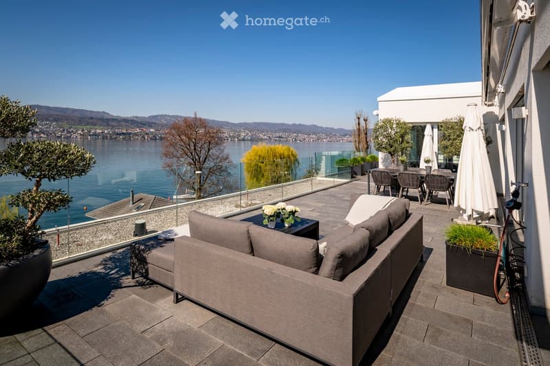 A modern outdoor terrace with a view of a lake and mountains, featuring a grey sofa, coffee table, dining area with chairs and table, and potted plants.