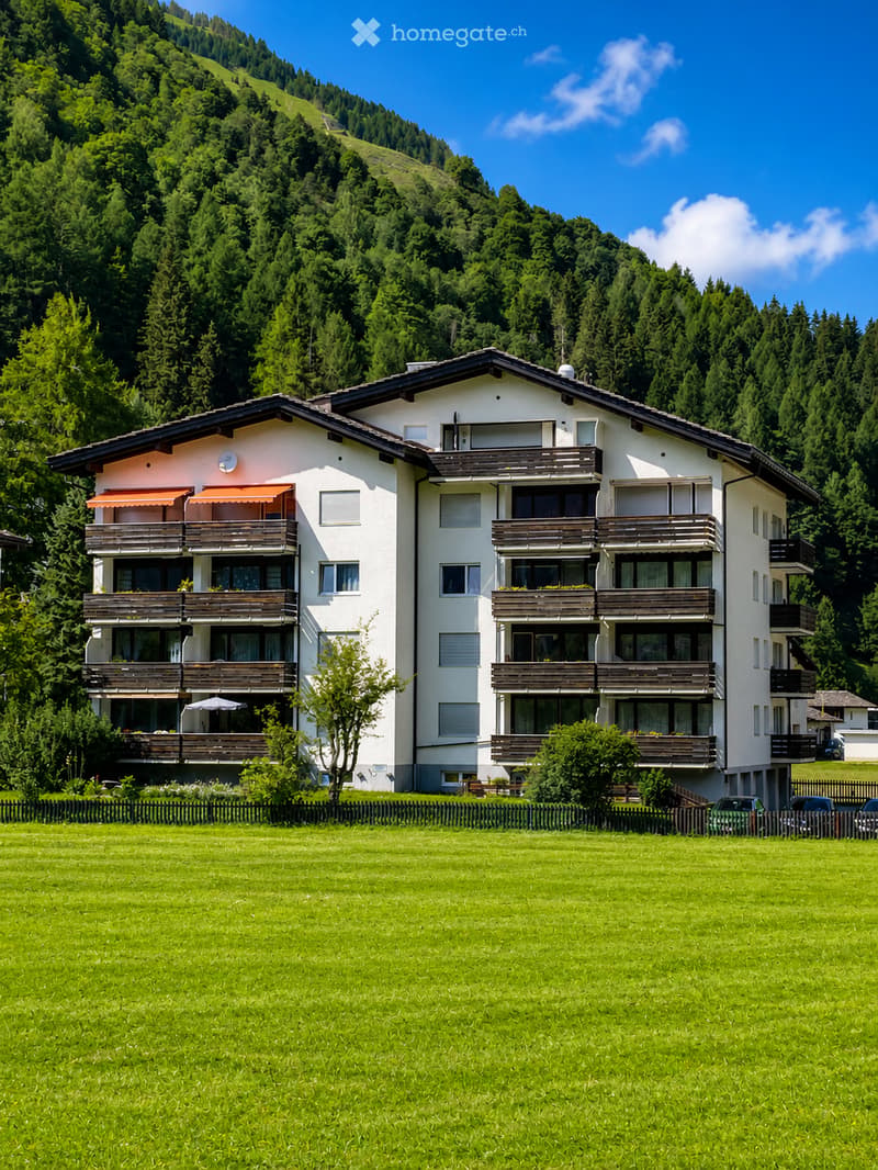 A two-story white building with multiple balconies and awnings, surrounded by a green lawn and trees, with a mountain view in the background.
