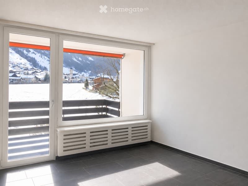 Empty room with large windows offering a scenic mountain view, white walls, dark tiled floor, radiator, and a balcony with black railings.