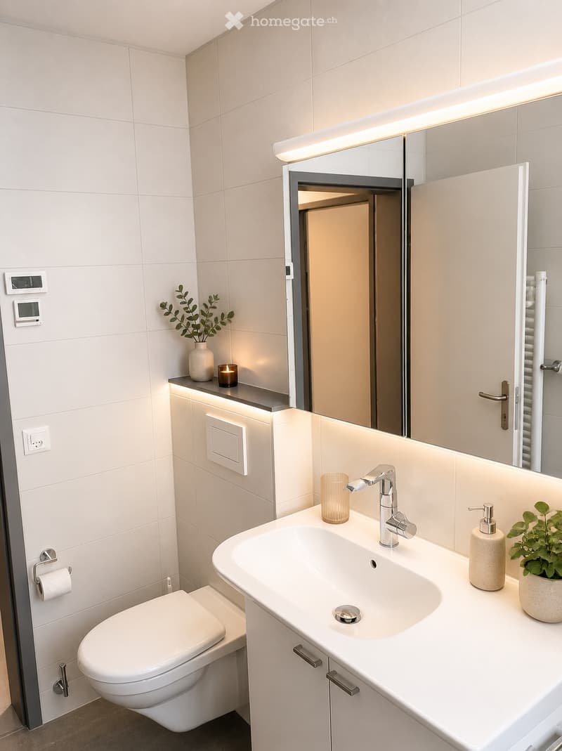 Modern bathroom with white tiles, a large sink with a chrome faucet, a toilet, a mirror with built-in lighting, and decorative plants and candles on the counter.