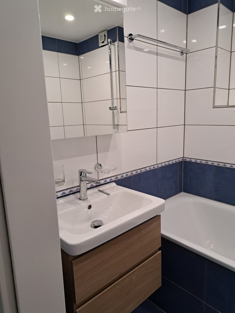 Modern bathroom with white tiles, blue accents, a white sink with a wooden cabinet, a chrome faucet, a bathtub, and a large mirror.