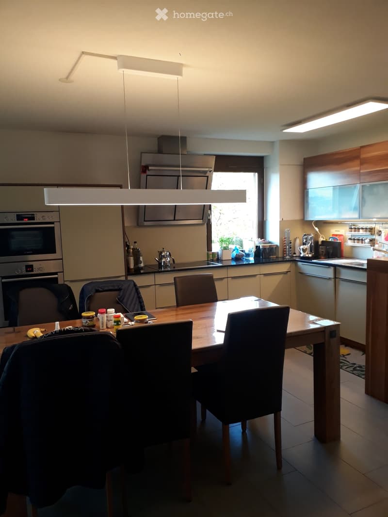 Modern kitchen with light-colored cabinets, stainless steel appliances, a wooden dining table with chairs, and various kitchen items on the counter.