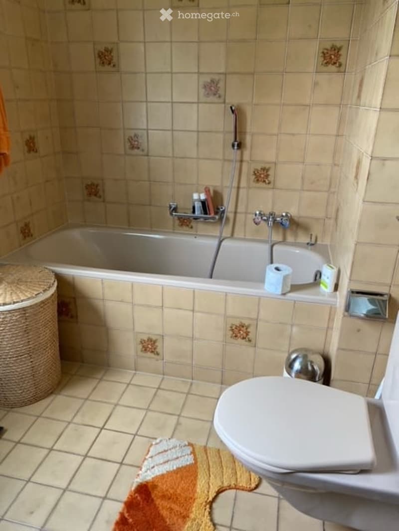 A small bathroom with beige tiled walls and floor, featuring a bathtub, toilet, shower hose, and a wicker basket on the floor.