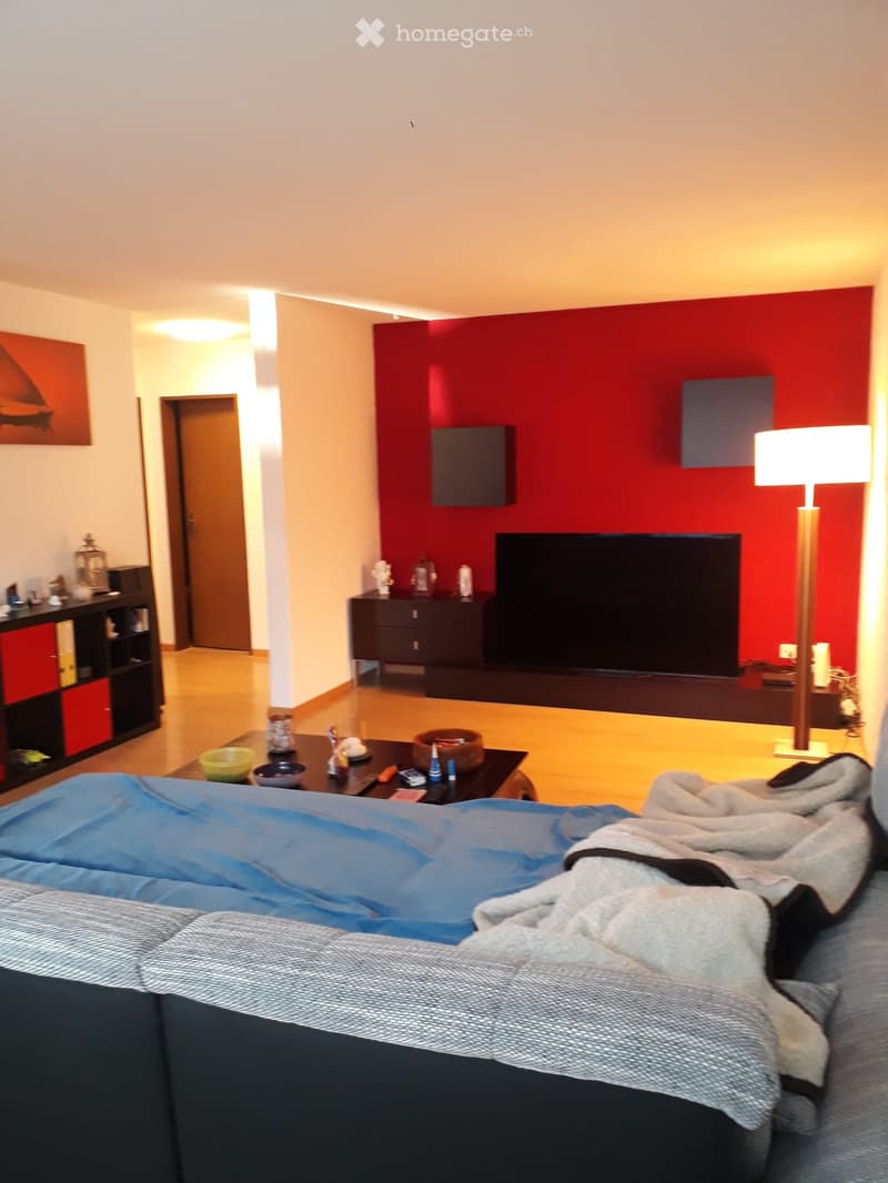 A modern living room with a red accent wall, a large flat-screen TV, a sofa with blankets, a coffee table with various items, a floor lamp, and a shelf with books and decorative items.