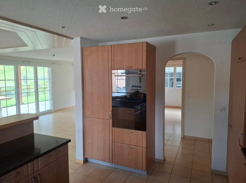 Modern kitchen with light wooden cabinets, built-in oven, and a black countertop. Large windows provide natural light, and the space is connected to an adjacent room through an arched doorway.