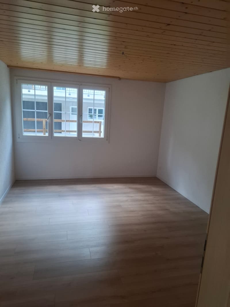 Empty room with wooden flooring, white walls, and large windows with white frames, allowing natural light to enter.