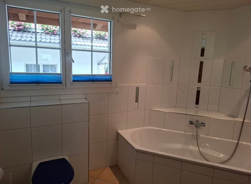 Modern bathroom with white tiled walls, a bathtub, toilet, and a large window with a view of a tiled roof and flowers outside.