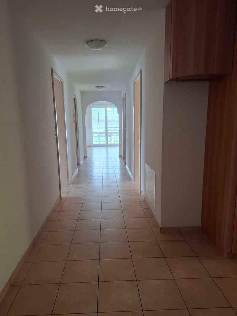 Empty hallway with tiled floor, beige walls, and several doors leading to other rooms. Natural light streams in from an arched window at the end.