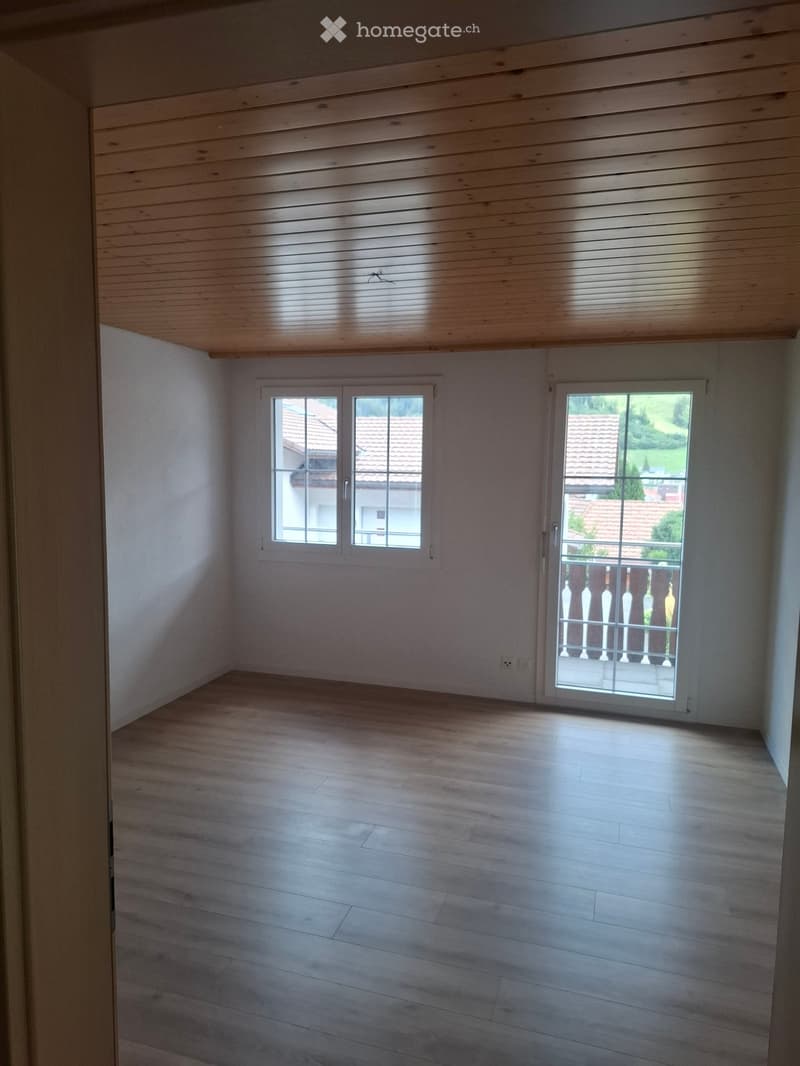 Empty room with wooden flooring, white walls, and large windows, including a sliding glass door leading to a balcony.