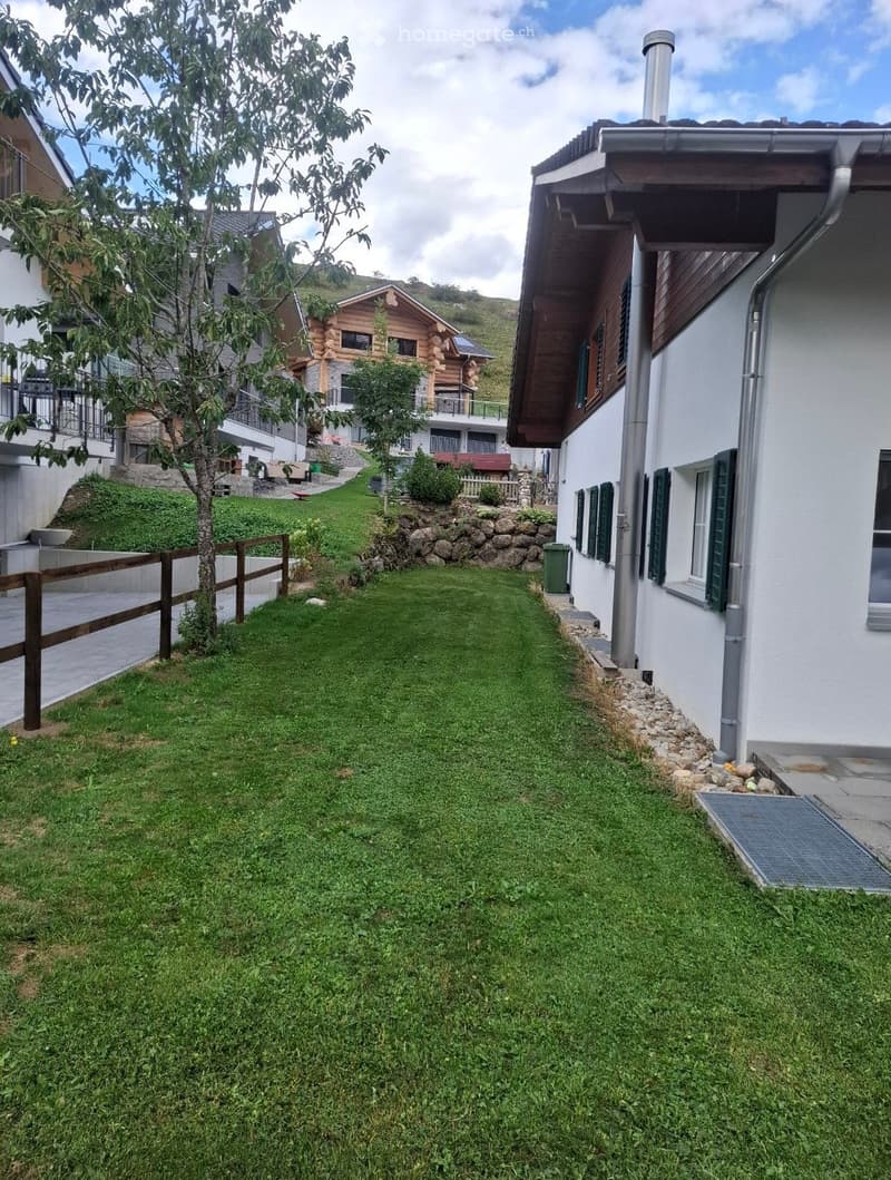 A well-maintained garden with a neatly trimmed lawn, a wooden fence, and a stone border along the house. The area is bordered by a white building on one side and a wooden fence on the other, with a small tree and shrubs adding greenery.