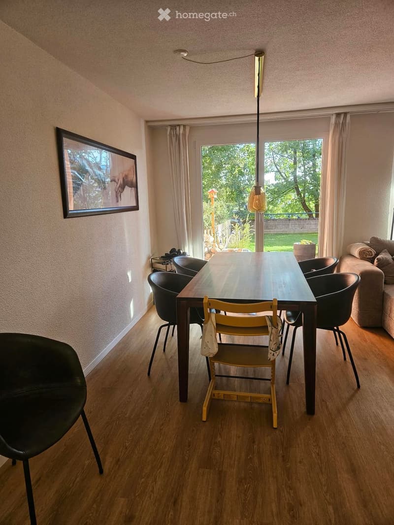 Dining area with a wooden dining table, black and yellow chairs, a sofa, and a picture frame on the wall. The room has wooden flooring and large windows with curtains, leading to a garden view.