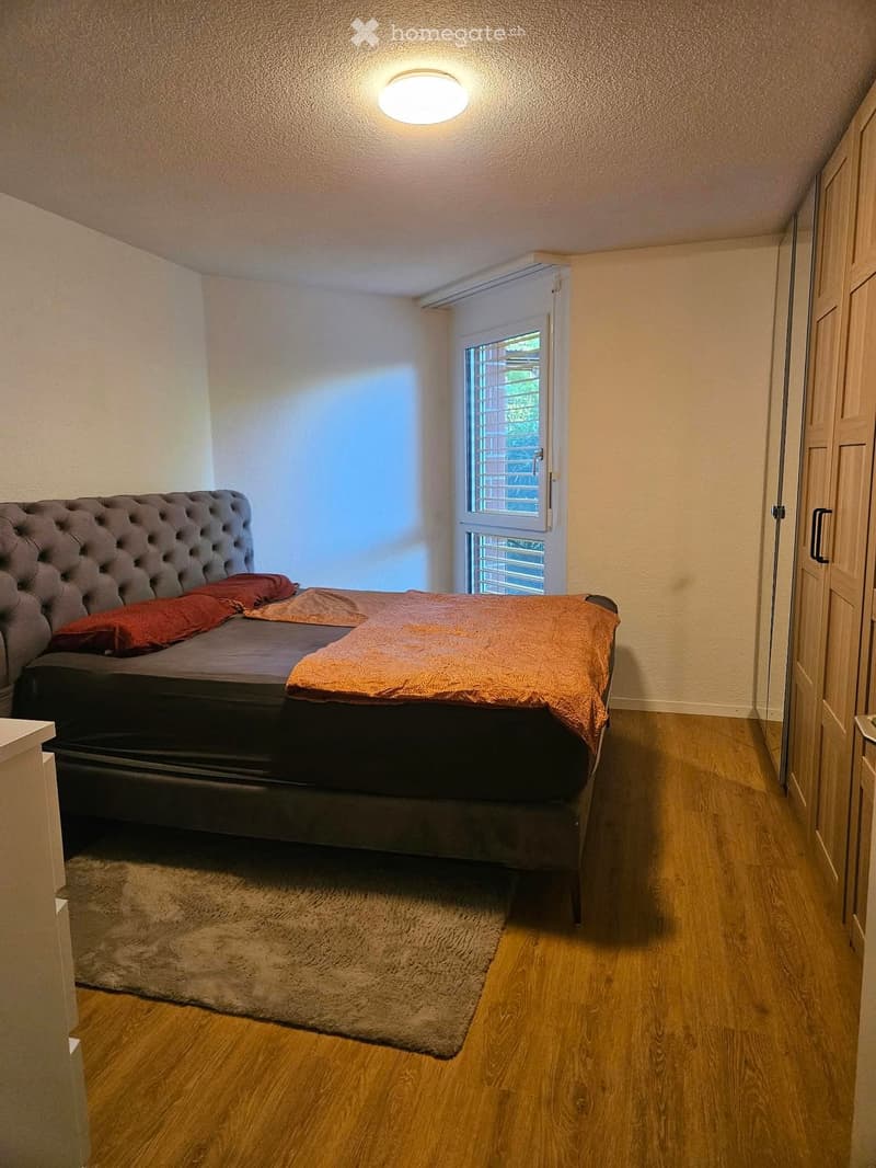 Cozy bedroom with a tufted headboard bed, orange blanket, pillows, and a soft rug on a wooden floor.