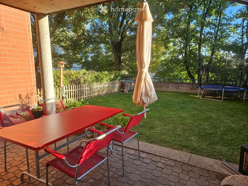 Outdoor patio with red table and chairs, umbrella, grassy lawn, wooden fence, and trampoline in the background.