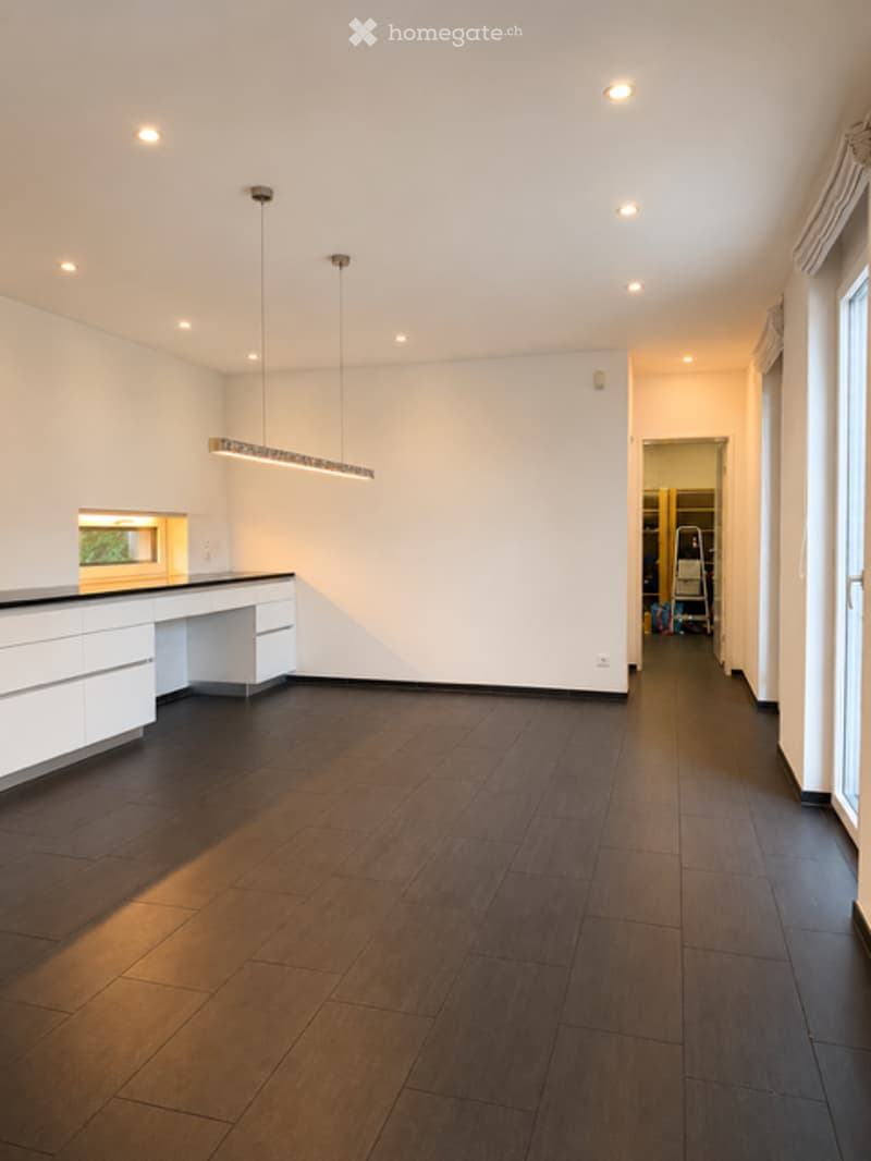 Completely empty room with white walls, dark wooden flooring, and recessed lighting. A small kitchen area is visible with white cabinets and a countertop.
