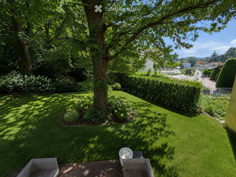 Well-maintained garden with a large tree, neatly trimmed hedges, and a small seating area with two wicker chairs and a table.