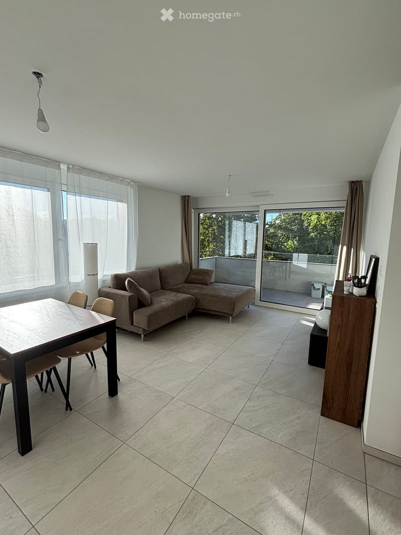Modern living room with a grey sectional sofa, a dining table with chairs, large windows with curtains, and a balcony access through glass doors.