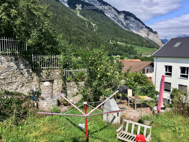 A serene garden area with a stone well, wooden bench, clothesline, and a small table surrounded by lush greenery and fruit trees, with a scenic mountain backdrop.