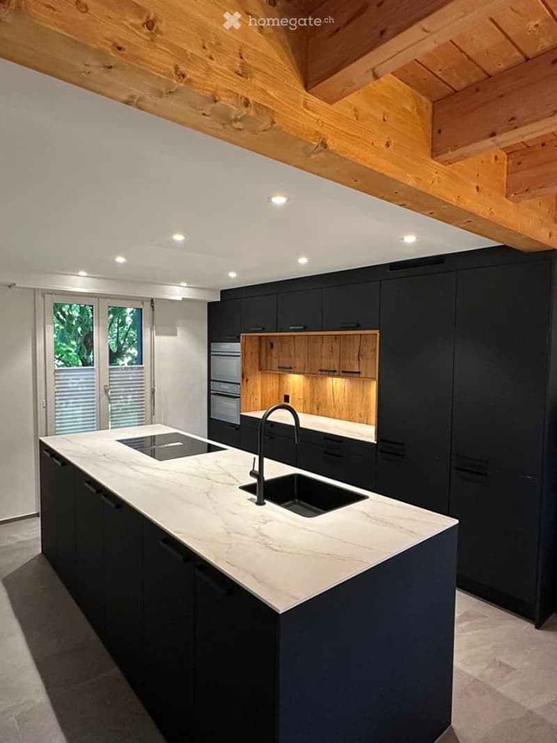Modern kitchen with a white marble island, black cabinets, and a stainless steel sink. Exposed wooden beams on the ceiling and recessed lighting.