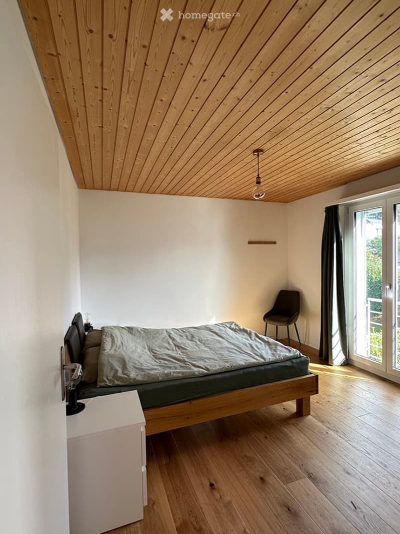 Cozy bedroom with wooden floors, slanted wooden ceiling, bed with grey duvet, nightstand, chair, and large window with curtains.