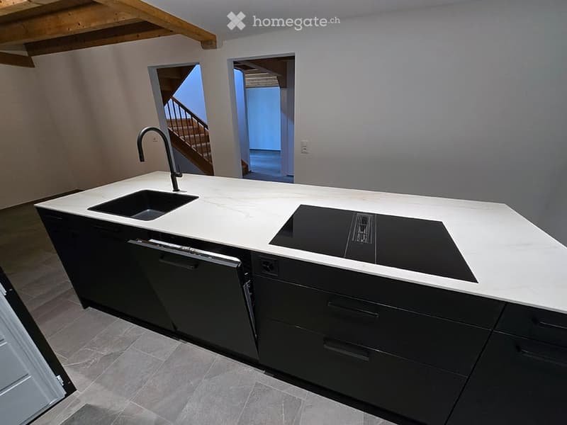 Modern kitchen with white countertops, black cabinets, a sink, faucet, dishwasher, and induction cooktop. The space features a visible staircase leading to another level.