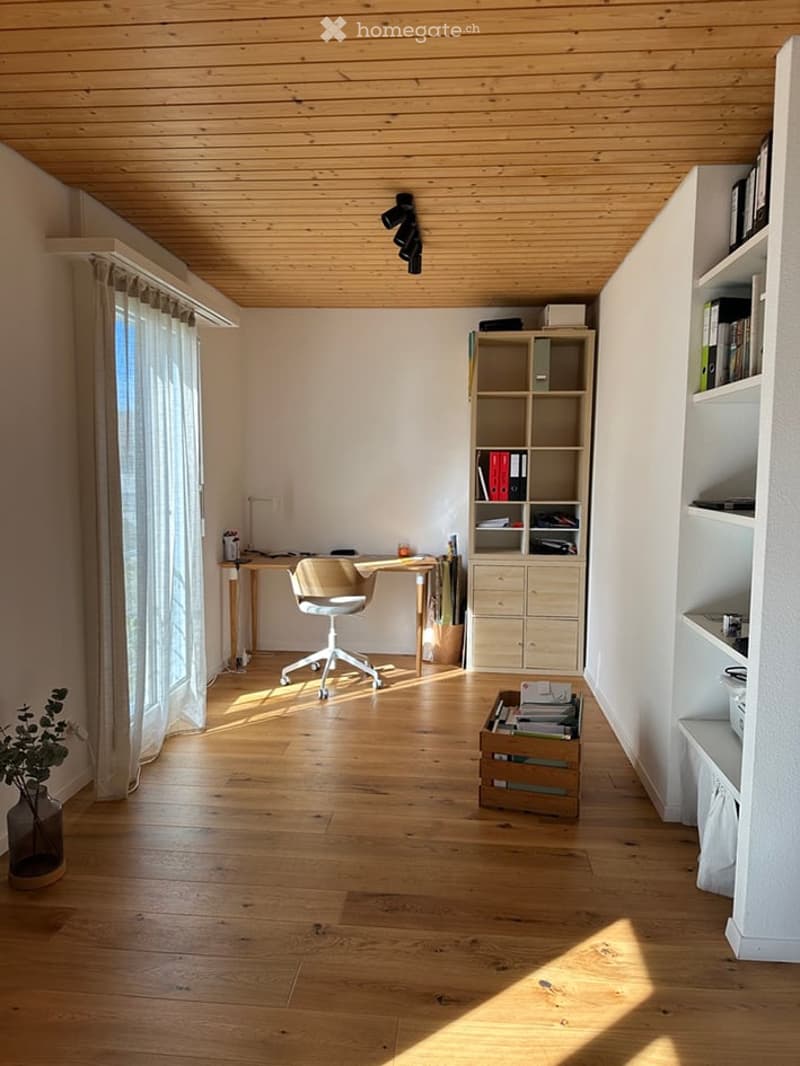 Bright, modern study or home office with wooden flooring, exposed wooden ceiling beams, large window with white curtains, desk with chair, bookshelf, and storage box.