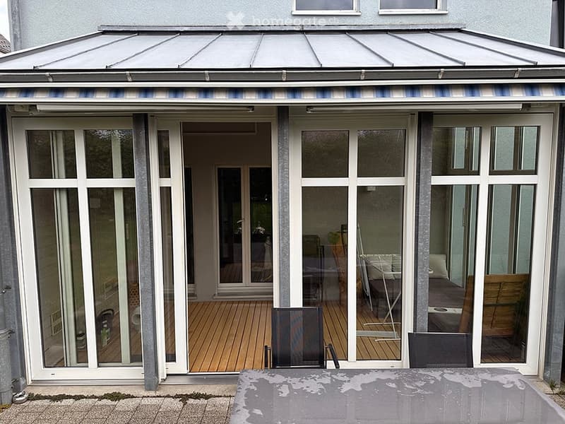 A covered patio with large glass doors leading to a wooden deck area inside the house. The patio has a retractable roof and is furnished with outdoor chairs and a table.