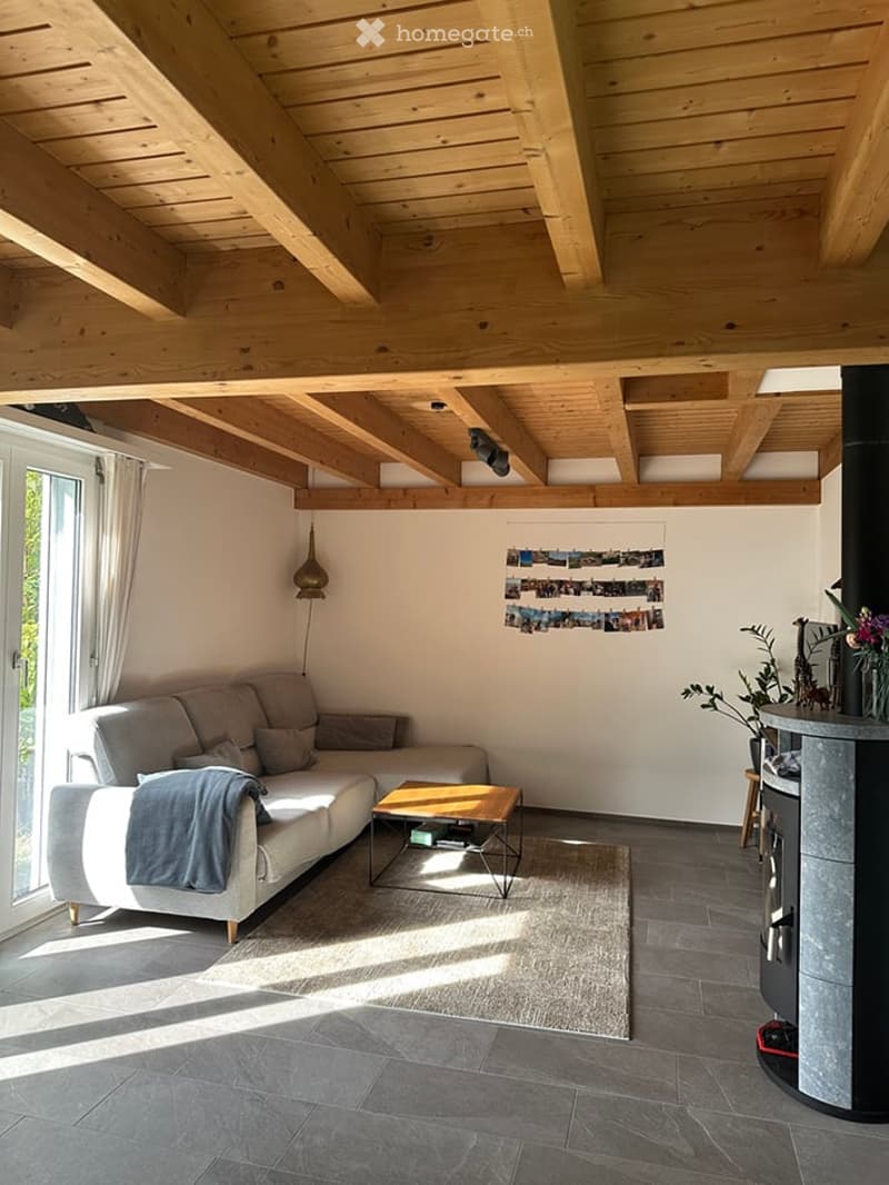 Modern living room with exposed wooden beams, grey sofa, coffee table, floor lamp, and a fireplace with decorative items on top.