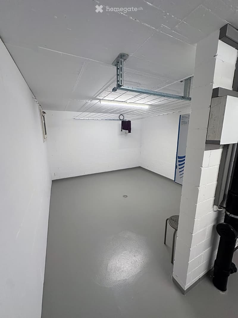 Empty room with white walls, grey floor, exposed pipes, and a small stool in the corner.
