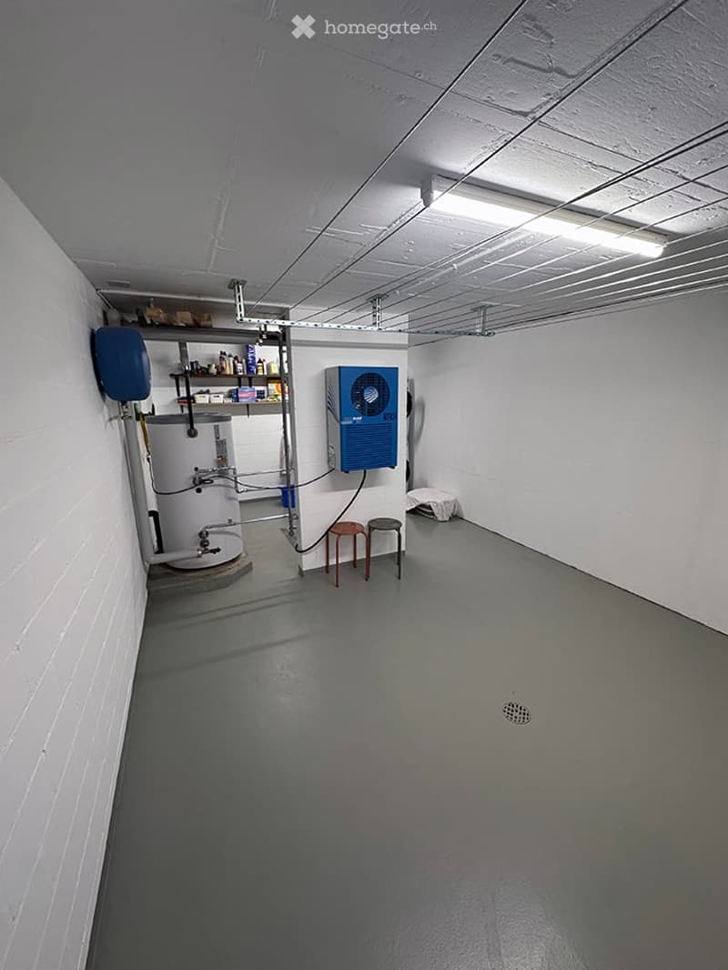 Empty utility room with white walls, grey concrete floor, exposed ceiling with lighting, water heater, air conditioning unit, shelving, and two stools.