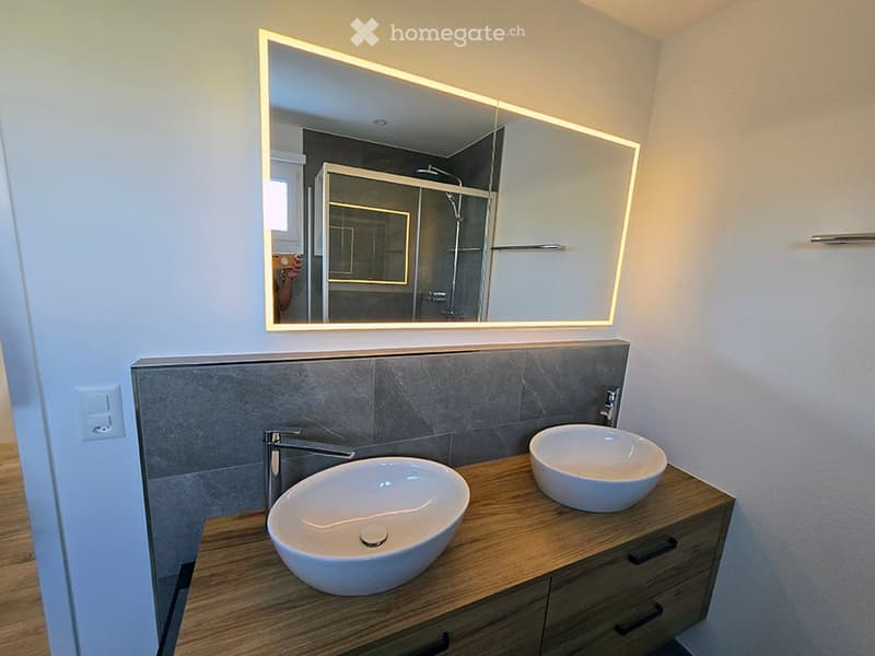 Modern bathroom with two white sinks on a wooden vanity, sleek faucets, and a large illuminated mirror above.